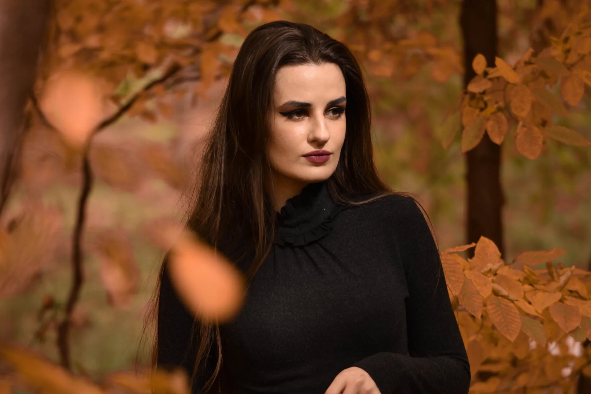 Młoda kobieta w czarnej sukience, pozująca w jesiennym lesie, otoczona liśćmi.